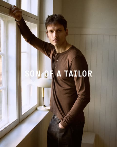A man stands next to a window wearing a long-sleeve shirt and jeans.