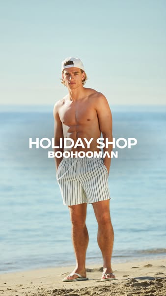 A man stands on a beach modeling swim trunks from boohooMAN's holiday shop.