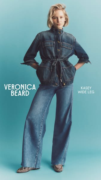 This image ad showcases a denim jacket and wide leg jeans from Veronica Beard. The model is posing in front of a solid b