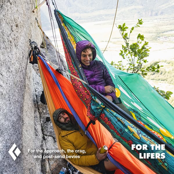 Two people are relaxing in hammocks attached to the side of a rock climbing face, with a mountain and valley visible in