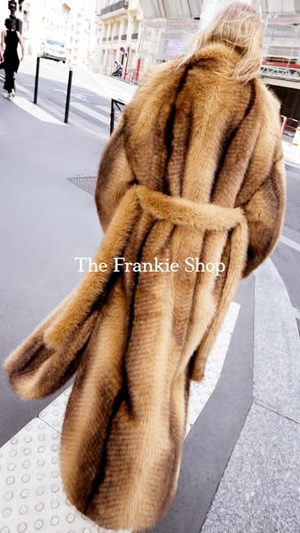 An image ad showcasing a brown fur coat from The Frankie Shop. The coat is worn on a city sidewalk, with the brand's