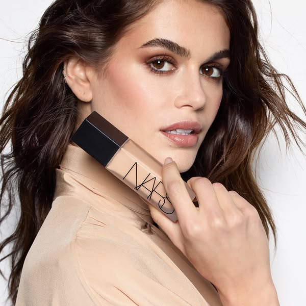 This image ad features model Kaia Gerber holding a NARS foundation bottle. The ad emphasizes the product's quality a