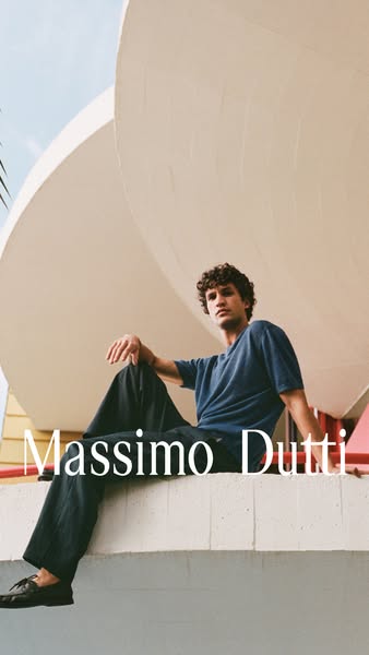 A man wearing Massimo Dutti sits on a ledge in front of a modern building.