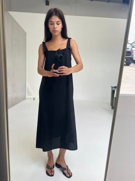 A woman takes a mirror selfie wearing a black dress and sandals.