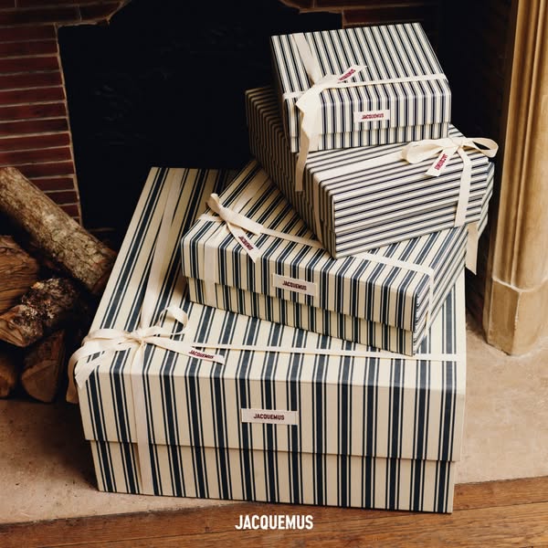 This ad features a stack of Jacquemus-branded gift boxes in front of a fireplace, suggesting a luxurious and cozy holida