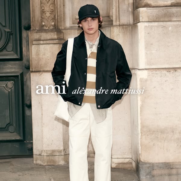 A person wearing a black jacket, beige and white striped sweater, white pants, and a black baseball cap in front of a st