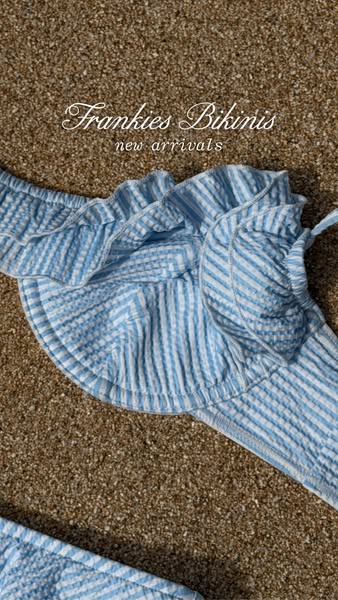 This image ad features a close-up shot of a blue and white striped bikini from Frankie's Bikinis, laid out on a sand