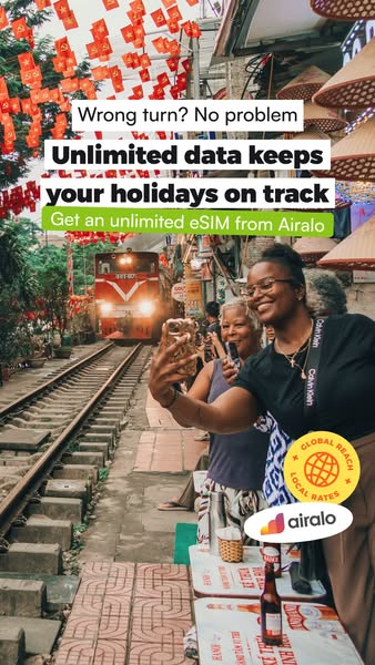This image ad for Airalo features two women taking a selfie on a bustling street with a train track running through it. 