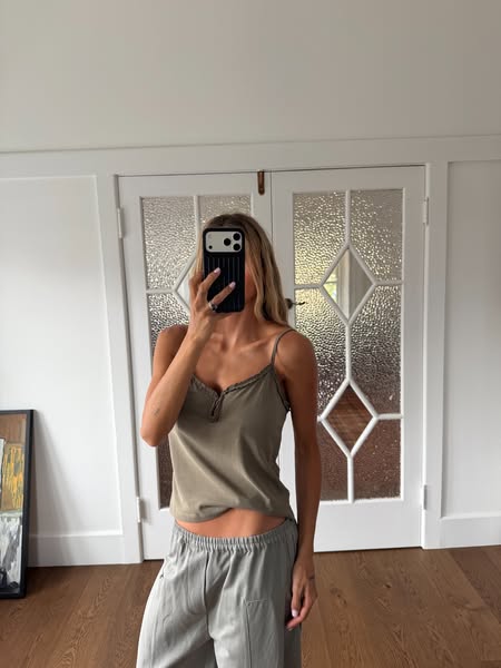 A woman takes a mirror selfie wearing an olive green camisole top and grey pants.