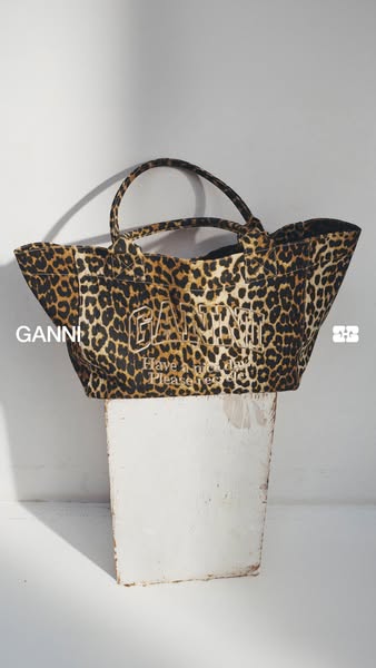 The ad features a GANNI brand leopard-print tote bag positioned on a white block. The bag is angled slightly towards the
