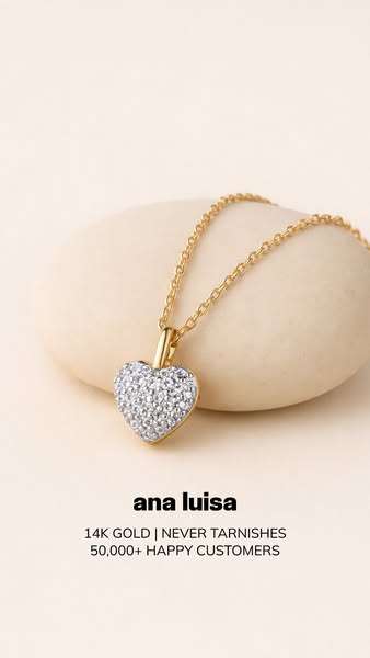 This ad features an Ana Luisa heart pendant necklace on a gold chain. The pendant is displayed resting on a neutral-colo