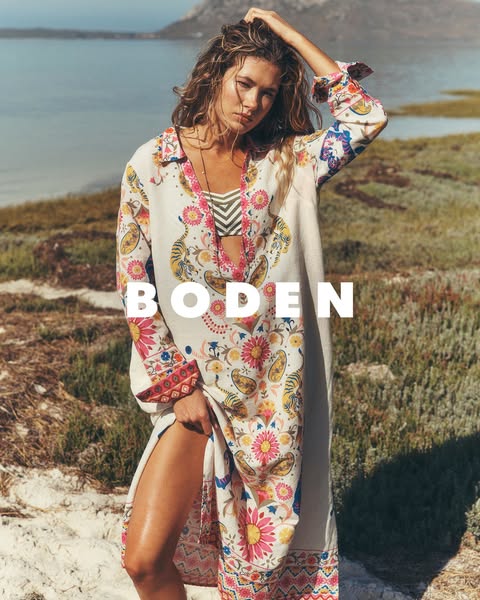 An ad for Boden features a woman standing on a grassy bank, looking at the camera and holding up the hem of a long patte