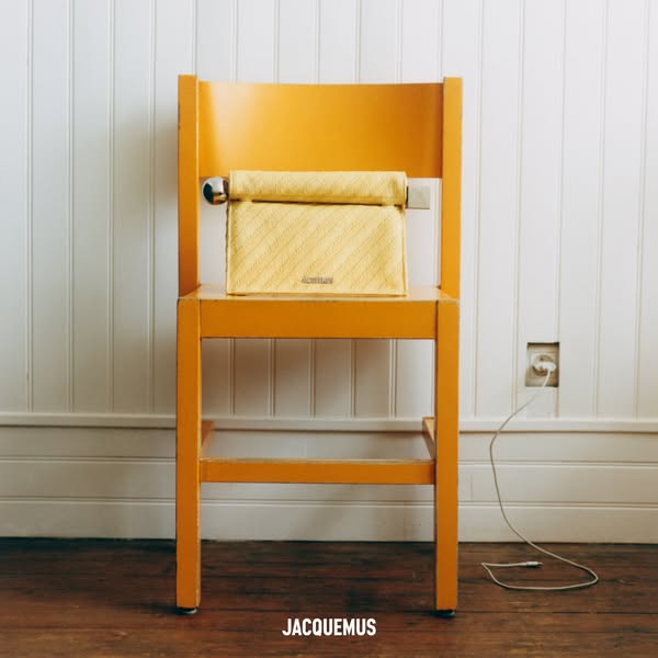 This image features a yellow Jacquemus handbag sitting on a yellow chair. An iPhone charger is plugged into the wall in 
