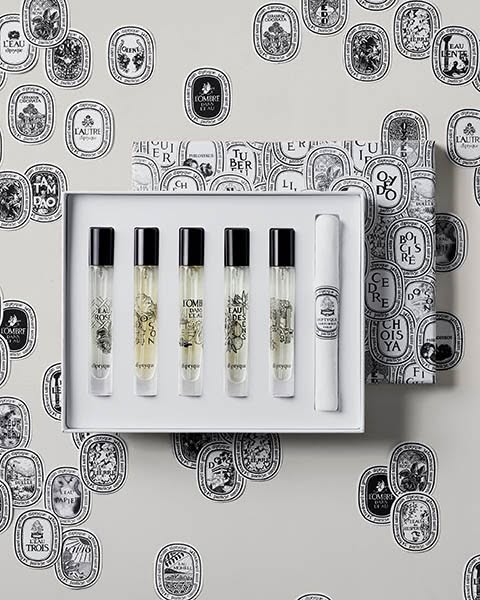 Ad showcasing the Diptyque perfume set against a background with the brand's oval logo repeated. The composition sug