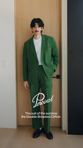 A stylish man in a green suit stands against a neutral wall to highlight the Percival Double-Breasted Clifton suit, whic