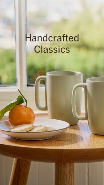 This ad features a lifestyle image showcasing handcrafted mugs on a wooden table with an orange and cookies. The focus i