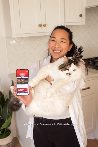 A woman holds her cat in one arm while displaying the Purina app on her phone in the other. The image promotes Purina an