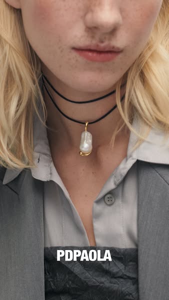 An image ad for PDPaola jewelry. It features a close-up of a woman wearing a black choker with a baroque pearl pendant f