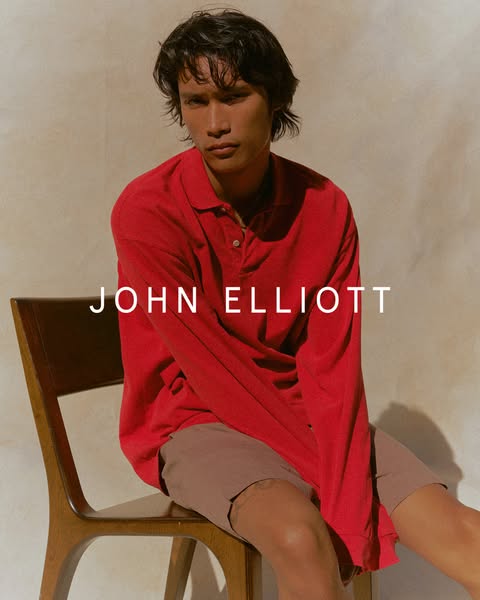 A man is seated and wearing a red long-sleeved polo shirt and brown shorts from designer John Elliott.