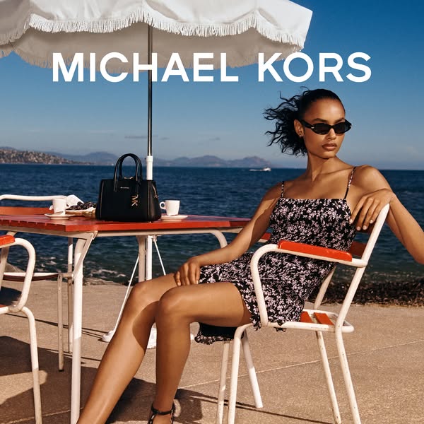 This ad features a woman sitting at a table on a patio overlooking the sea, with a Michael Kors handbag on the table nex