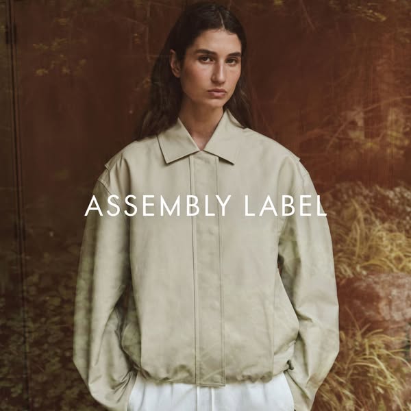 This ad features a woman wearing a light-colored jacket over white pants. The brand name, ASSEMBLY LABEL, is displayed i
