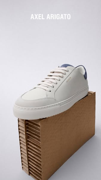 A product ad showcasing a white Axel Arigato sneaker on a brown cardboard box against a white background.