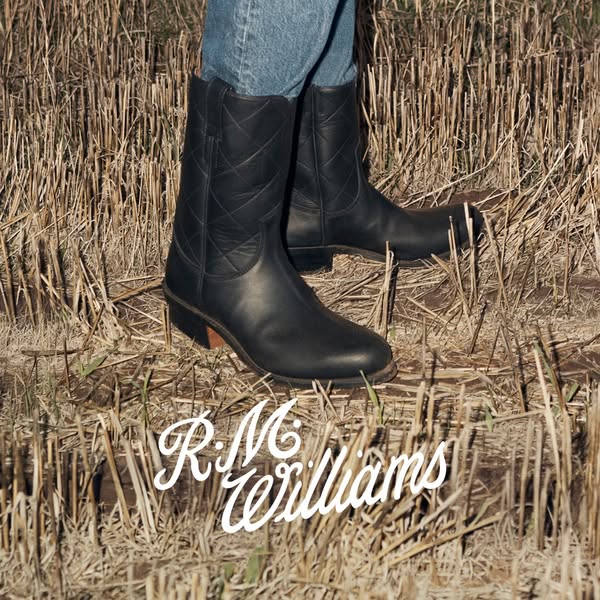 This ad showcases a pair of R.M. Williams boots in a field of dry grass. The brand logo is featured in a stylized script