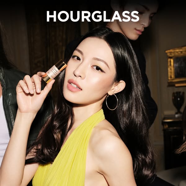 An ad featuring a woman holding an Hourglass foundation stick. The background is staged to look like a social gathering.