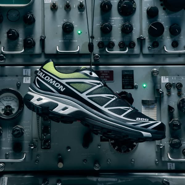 This ad showcases a black Salomon athletic shoe. The shoe is suspended on a string in front of a panel of grey electroni