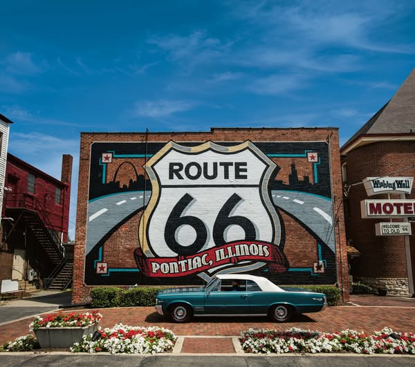 This image depicts a teal vintage convertible parked in front of a mural of the Route 66 sign on a brick building in Pon