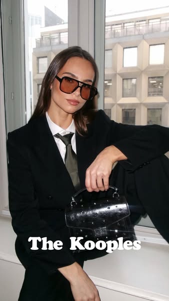 This image ad by The Kooples features a model in a power suit and sunglasses, holding a transparent purse with studs. Th