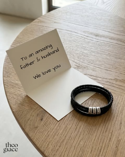 The ad features a black braided bracelet with three engraved charms on a wooden table next to a card that reads, “To an