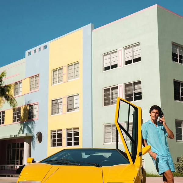 A man in a matching blue outfit stands next to a yellow Lamborghini in front of a pastel-colored building.