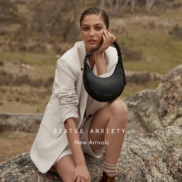 This ad for Status Anxiety showcases a woman sitting outdoors on a rock, modeling one of the brand's black handbags.