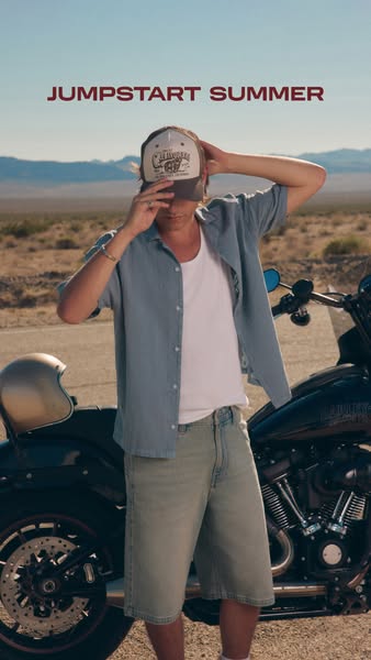 A male model stands next to a motorcycle in an outdoor setting, wearing a hat, t-shirt, shirt, and shorts, and the words