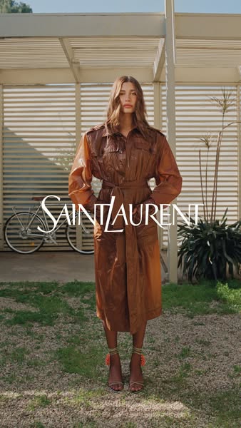 This Saint Laurent ad features a model in a brown leather trench coat with sandals, set against a neutral background wit