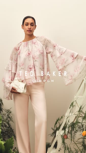 A woman models a floral print blouse and light pink pants from Ted Baker London, holding a white clutch.