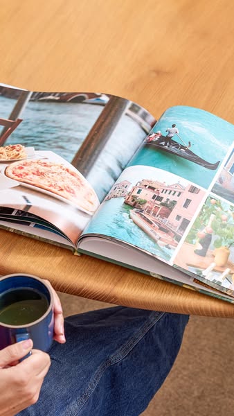 This ad features an open photo album on a wooden surface. A woman's hands are visible holding a mug. The album conta