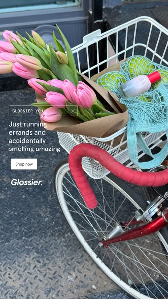 The image shows a bicycle with a basket that contains a bouquet of tulips, green fruit, and a Glossier product, with the