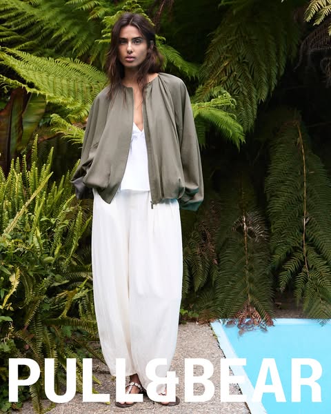 This ad features a woman showcasing the brand's light green bomber jacket and white pants near a pool and lush green