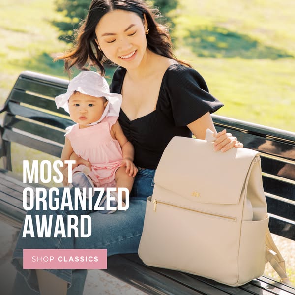 Image ad for a diaper bag, showing a mother holding her baby while sitting on a park bench with the bag next to her. The