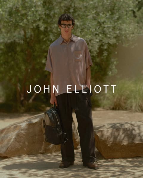This ad features a man standing in front of a natural outdoor background, showcasing clothing from the brand John Elliot