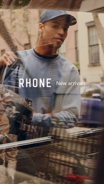 This image ad presents Rhone apparel. The visual is of a man wearing a blue shirt and a baseball cap, set against an urb