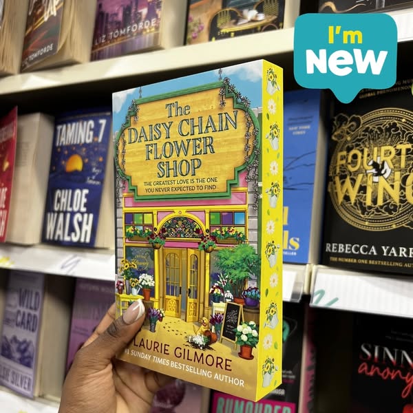 This ad features a hand holding a paperback book called 'The Daisy Chain Flower Shop'. The book is presented as 
