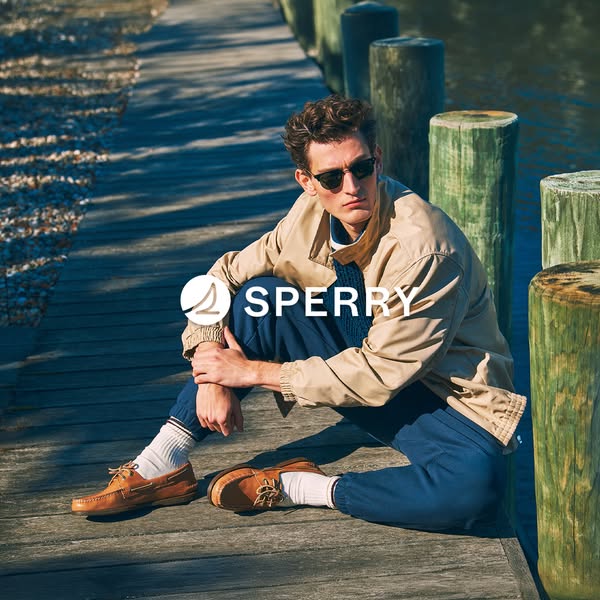 A man in Sperry shoes sits on a boardwalk and looks off into the distance.