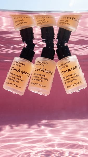 This image ad features three bottles of Champo Pitta Growth Serum suspended in clear water with a pink background. The s