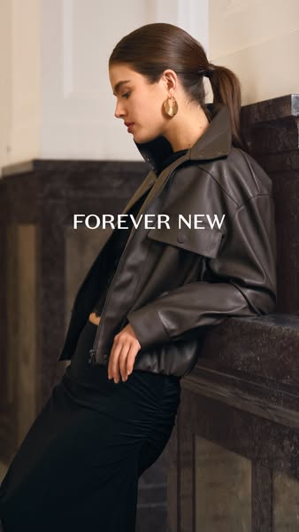 This image ad features a brunette woman leaning against a marble pillar wearing a dark brown jacket and a black dress. T