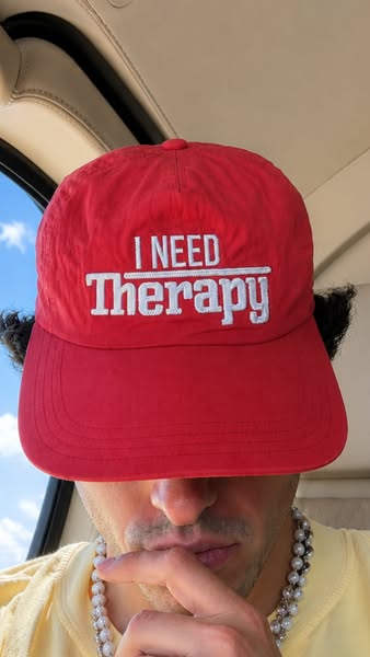 The ad is a selfie-style shot of a person wearing a red baseball cap with the phrase "I NEED Therapy" embroide
