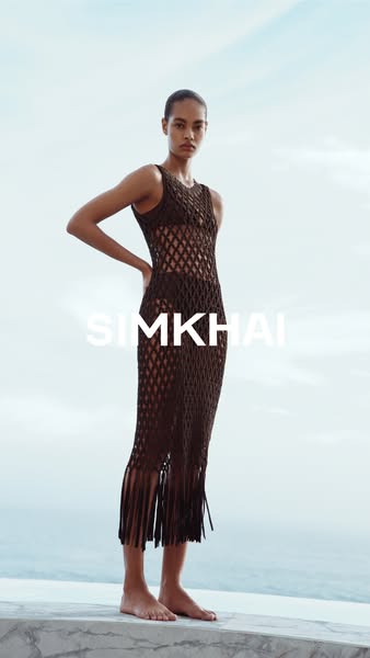 This ad showcases a SIMKHAI dress worn by a model standing against a bright outdoor background of water and sky. The sho