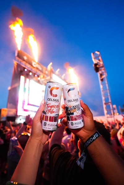 This image shows two Celsius energy drinks being held up at a music event. The ad is visually busy with a crowd of peopl